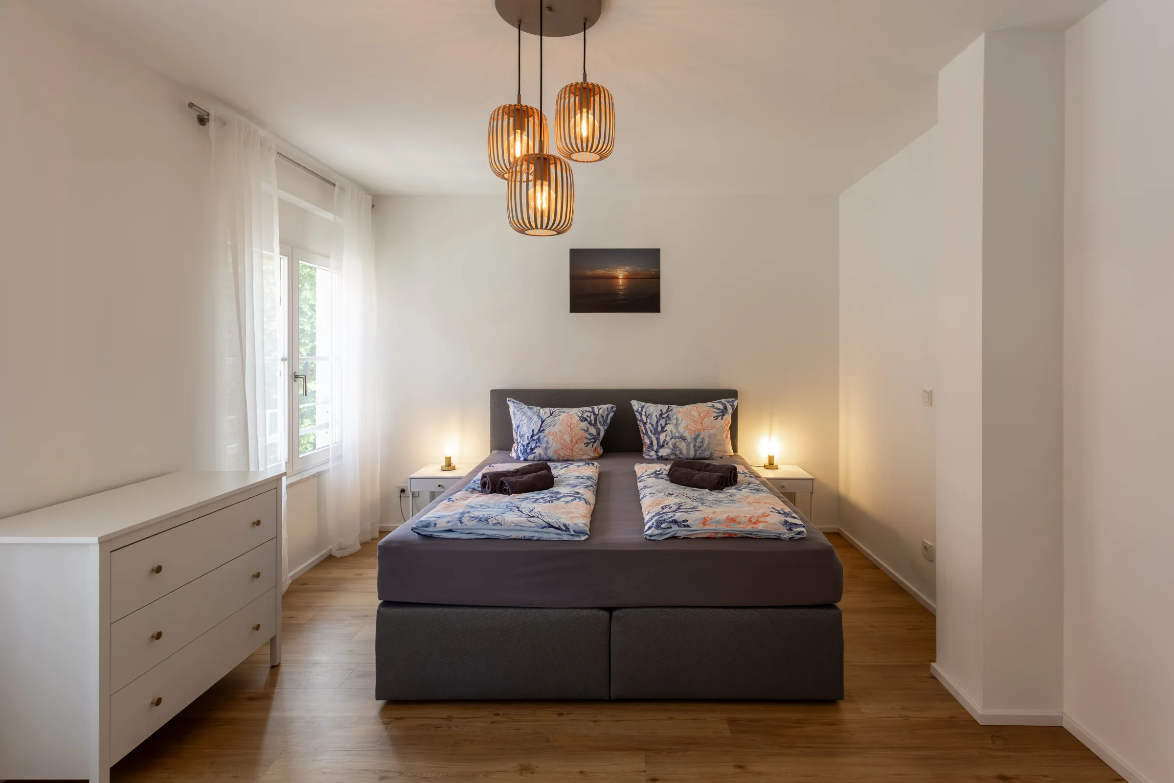 Hell eingerichtetes Schlafzimmer mit Doppelbett, gemusterter Bettwäsche, drei hängenden Holzpendelleuchten und Nachttischen