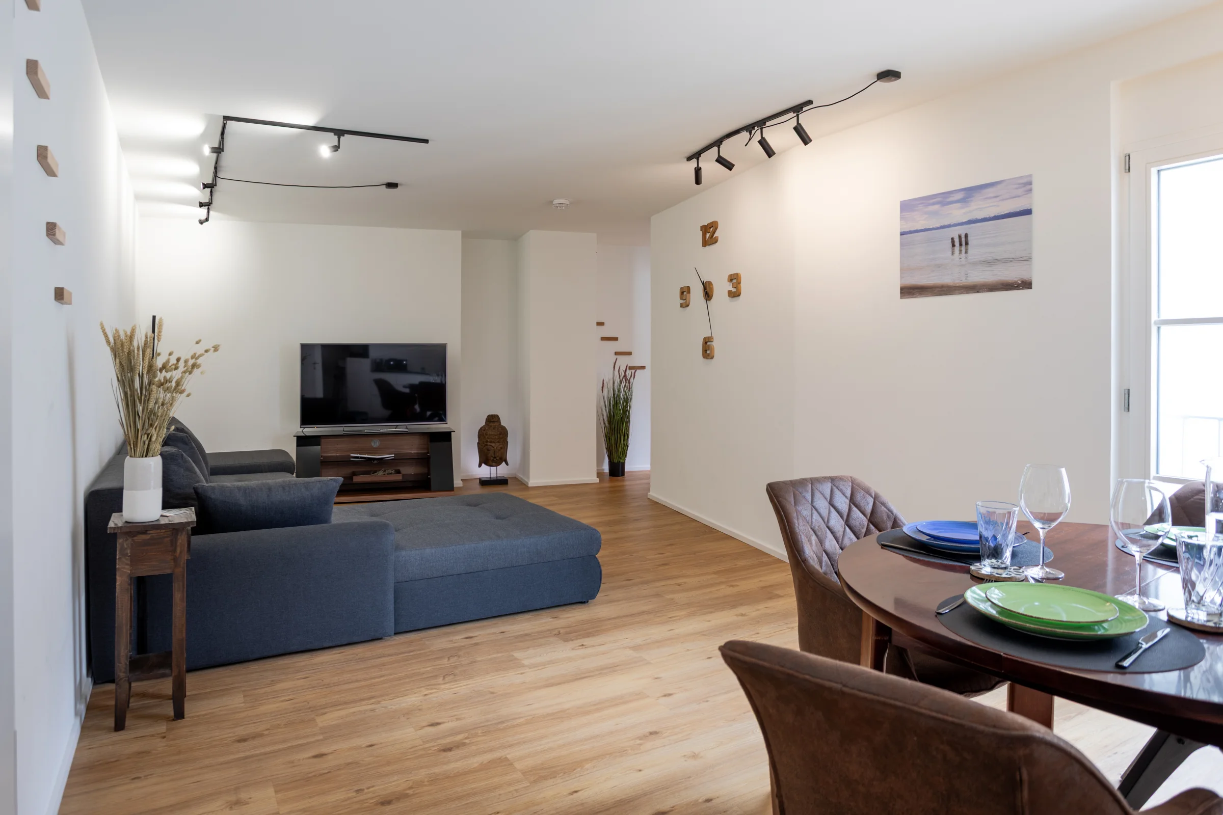 Offener Wohnbereich mit grauem Ecksofa, TV, Holzboden, Esstisch und Wanduhr mit Holz-Zahlen