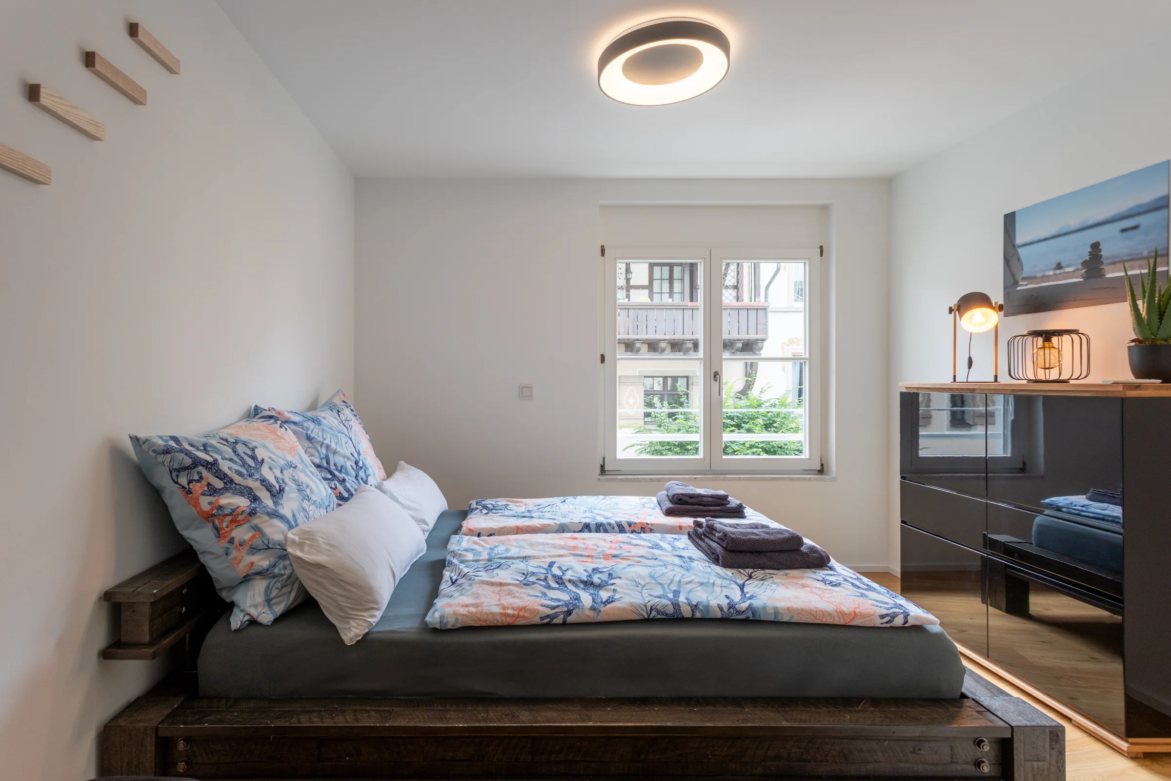 Helles Schlafzimmer mit Doppelbett, blau-oranger Bettwäsche, Fenster, schwarzem Sideboard und zwei dekorativen Lampen