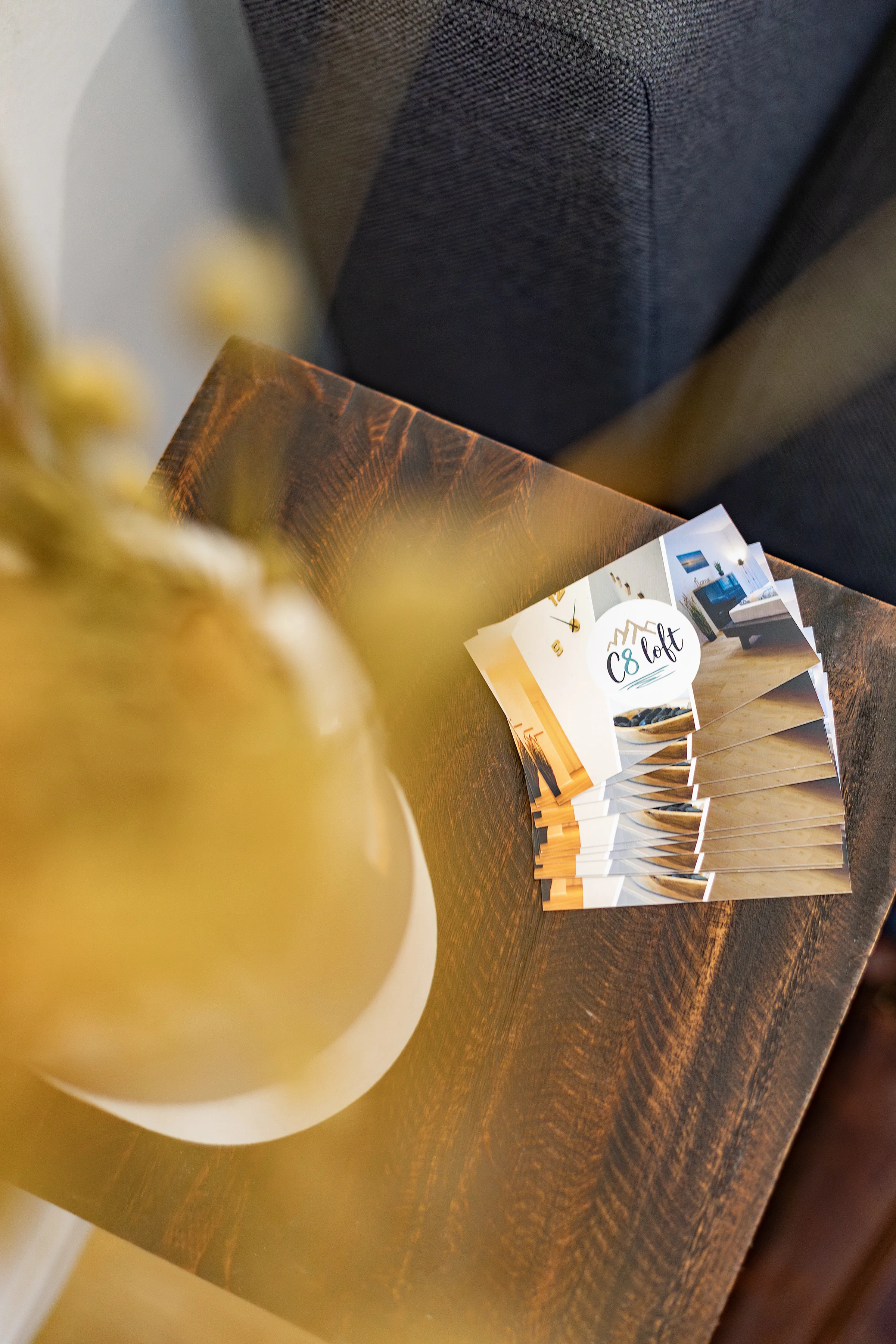 Gestapelte C8 Loft‑Flyer auf dunklem Holztisch, graues Sofa im Hintergrund, unscharfe goldene Lampe im Vordergrund
