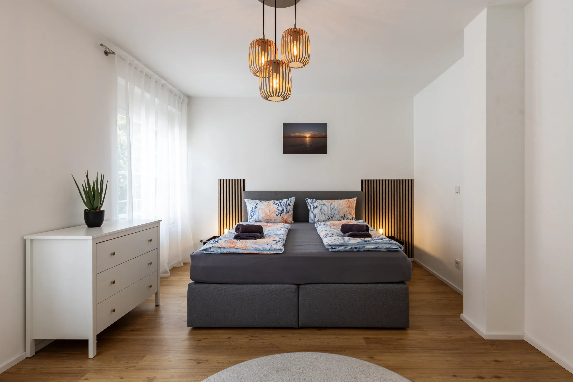 Symmetrisches Schlafzimmer mit grauem Bett, zwei gemusterten Decken, hängenden Holzlampen und weißer Kommode
