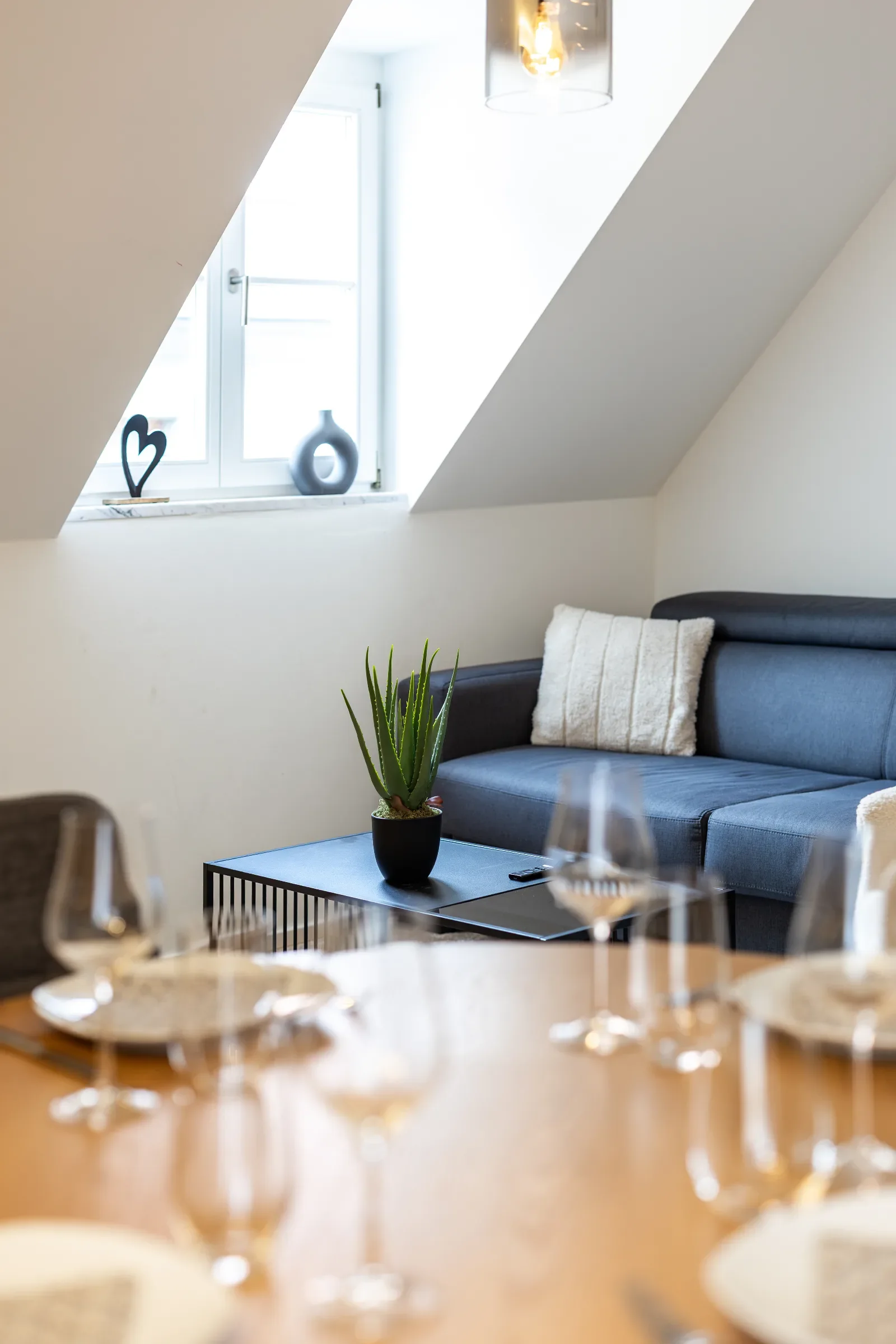 Sitzecke mit dunkelgrauem Sofa, hellem Kissen, Couchtisch mit Topfpflanze und dekoriertem Fenster unter Dachschräge