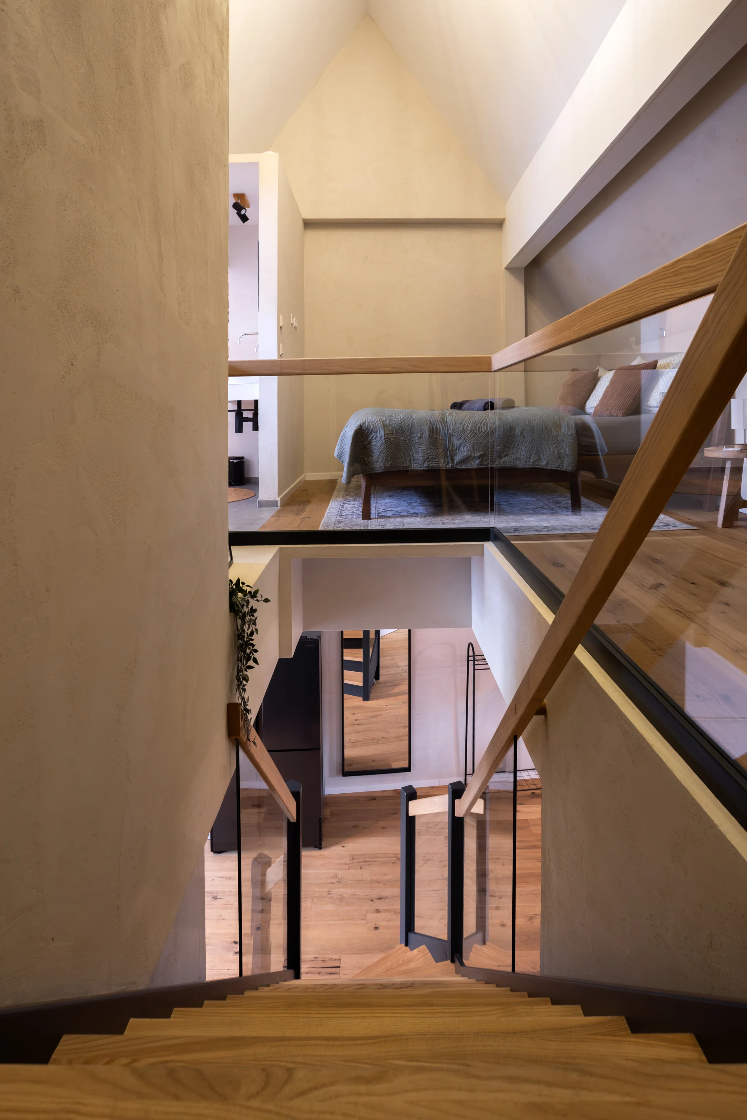 Galerie mit Holztreppe und Glasgeländer, Blick auf ein Bett mit gesteppter Decke und Teppich
