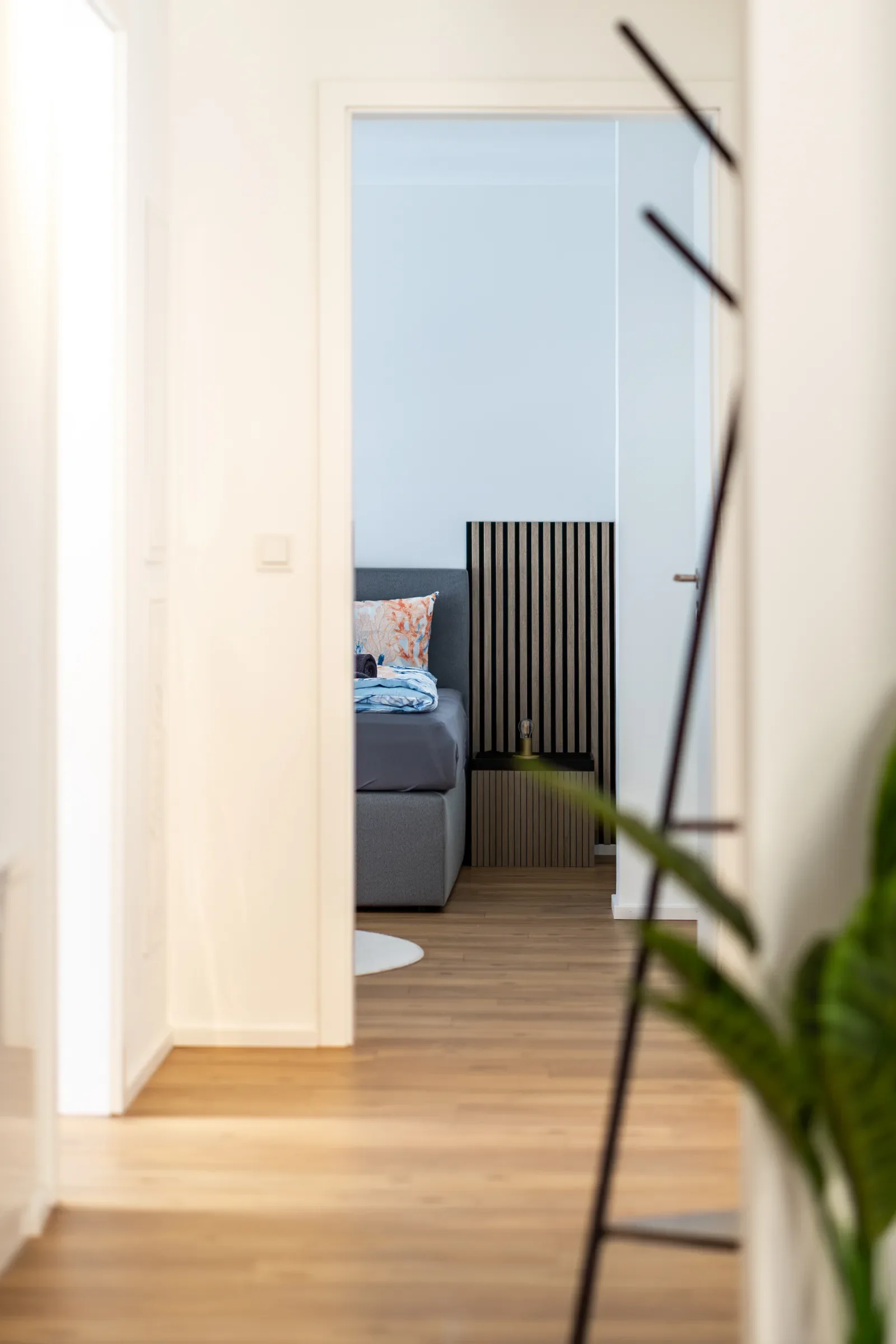 Blick durch einen Flur in ein Schlafzimmer mit grauem Bett, Nachttisch mit vertikalen Lamellen und kleiner Lampe
