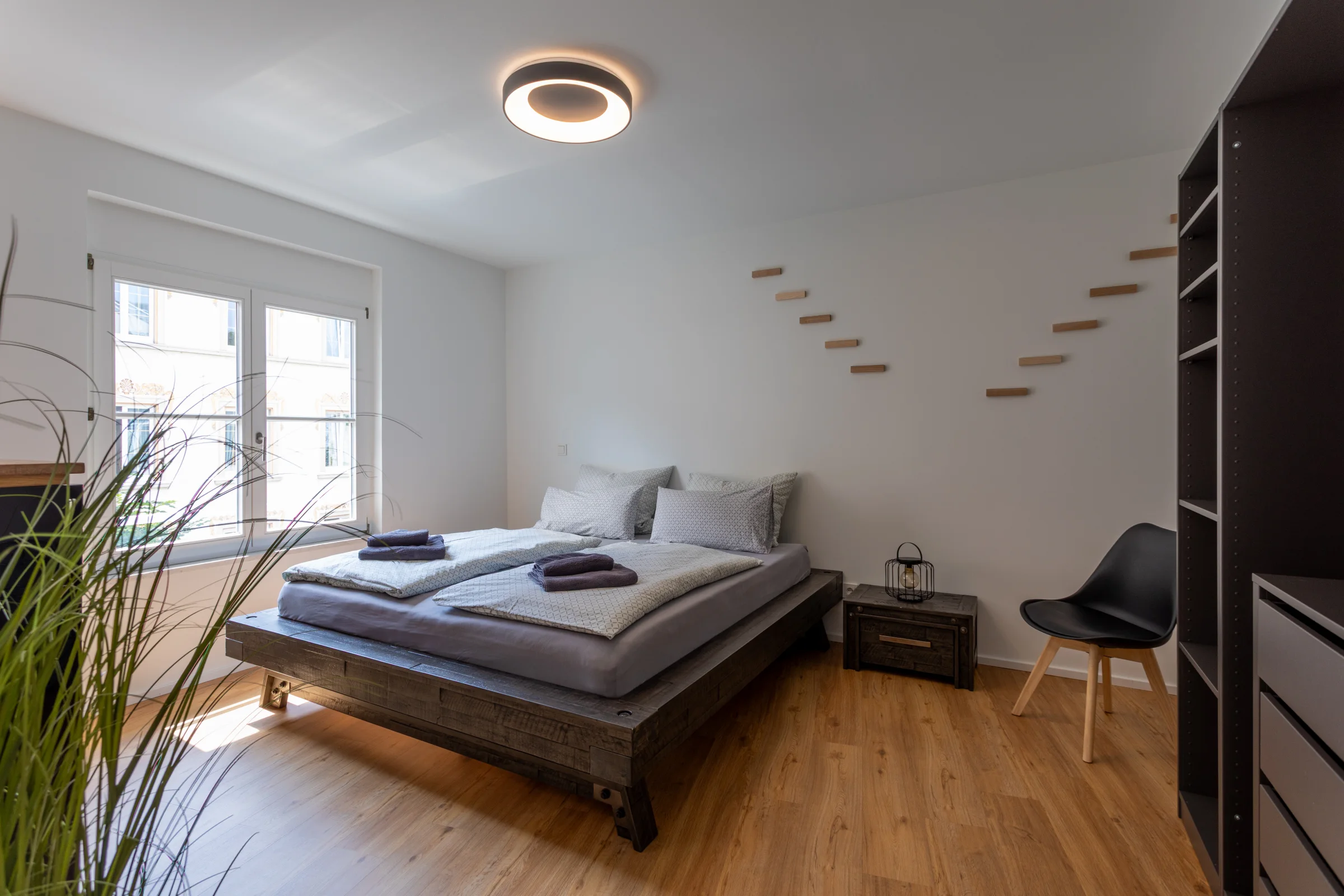 Helles Schlafzimmer mit niedriger Holzplattform, zwei Matratzen, Fenster, Deckenleuchte und Zimmerpflanze