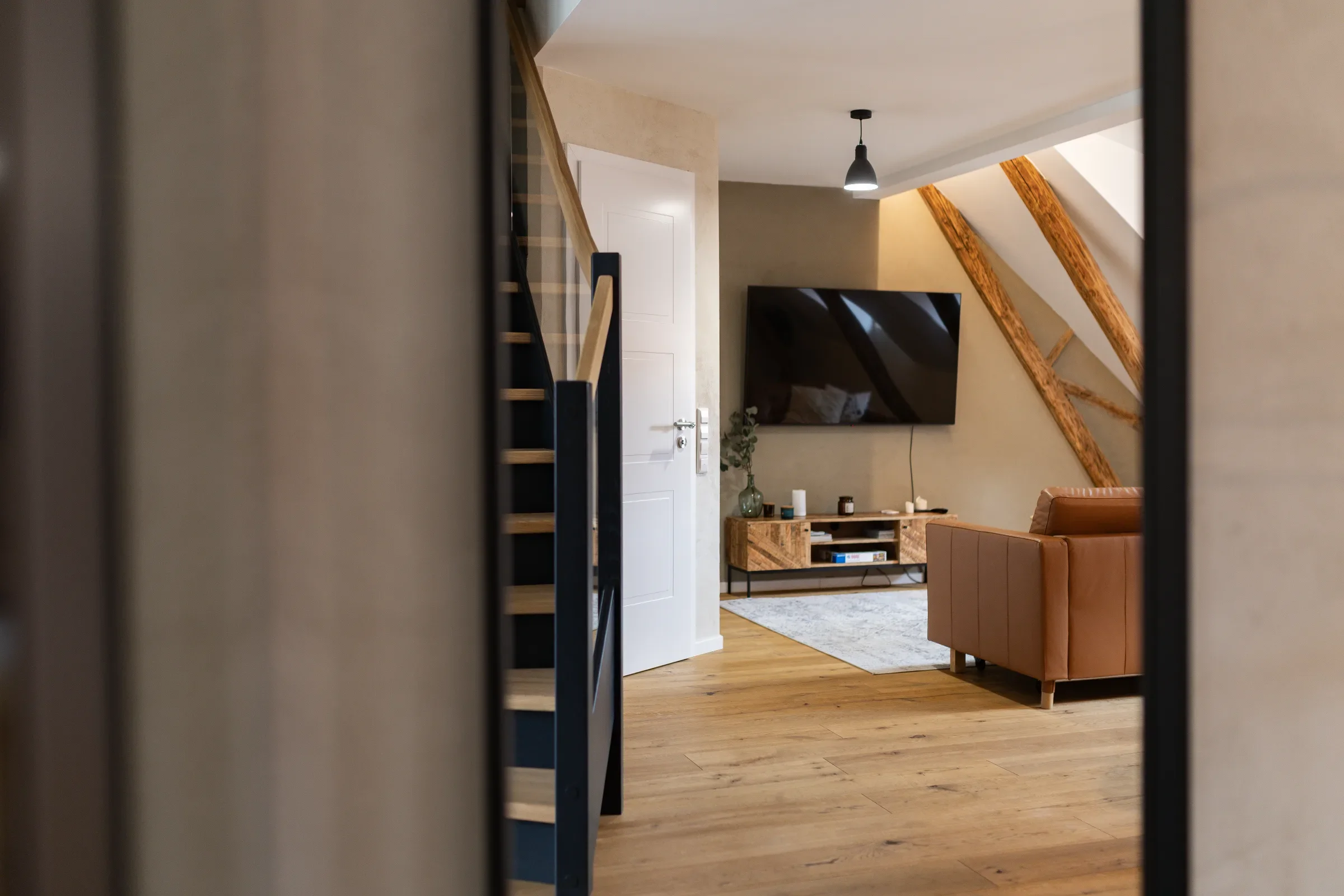 Offener Wohnbereich mit Holztreppe, wandmontiertem Fernseher, sichtbaren Holzbalken und brauner Ledercouch